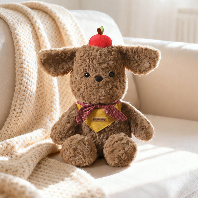 A high-quality brown plush dog wearing a yellow plaid bandana and a red apple on its head, sitting on a textured cream blanket in warm sunlight, showcasing premium OEM manufacturing quality, intricate stitching, and factory-direct production capabilities for global wholesale buyers.