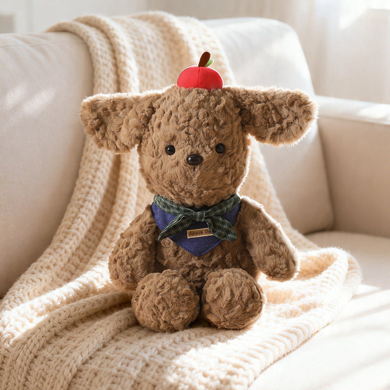 A sophisticated brown peluche (plush dog) wearing a subdued navy-blue plaid bandana and a red apple, sitting on a textured throw blanket. The low-saturation color palette and refined craftsmanship perfectly illustrate the "Quiet Design" trend and high-end quality demanded by the European and French wholesale markets.